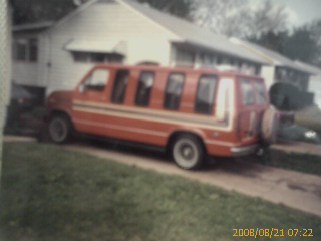 Orange van with multiple windows and spare tire on rear parked in front of a house.