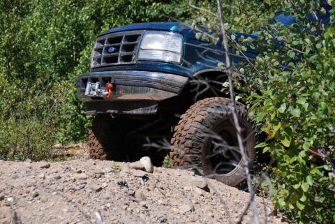 1996 Ford Bronco - A teal Ford Bronco with off-road tires is seen partially hidden by trees and brush on a rocky incline.