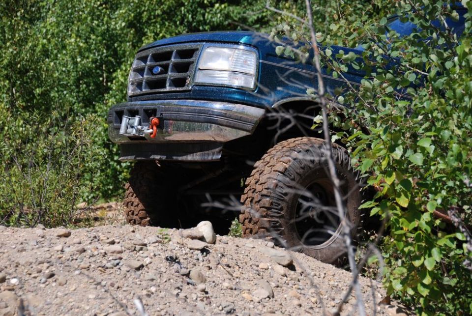 Teal Ford Bronco with winch on front bumper, off-road tires, partially obscured by foliage