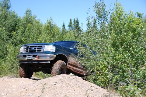1996 Ford Bronco - A dark-colored SUV navigates a sandy dirt road surrounded by lush green trees.