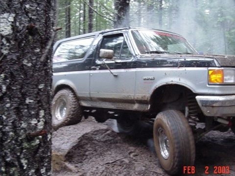 1989 Ford Bronco - A two-tone Ford Bronco is shown off-roading through a wooded area with muddy ground and large rocks.