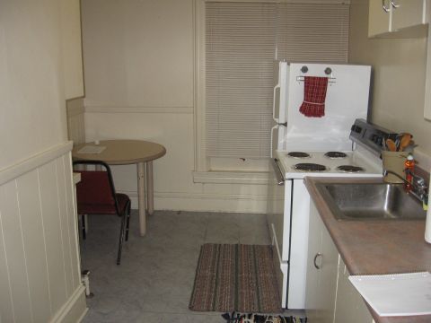 1984 Ford Bronco - A small kitchen area with a round table and chair, a stove, and a sink. A red and white checkered towel hangs on the refrigerator door.