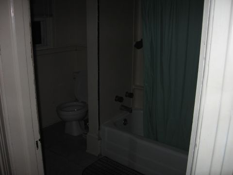 1984 Ford Bronco - A dimly lit bathroom contains a toilet and a bathtub with a green shower curtain. The image is taken from a doorway looking into the room.