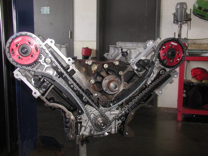 V8 engine block with timing chains and red cam gears, mounted on a stand