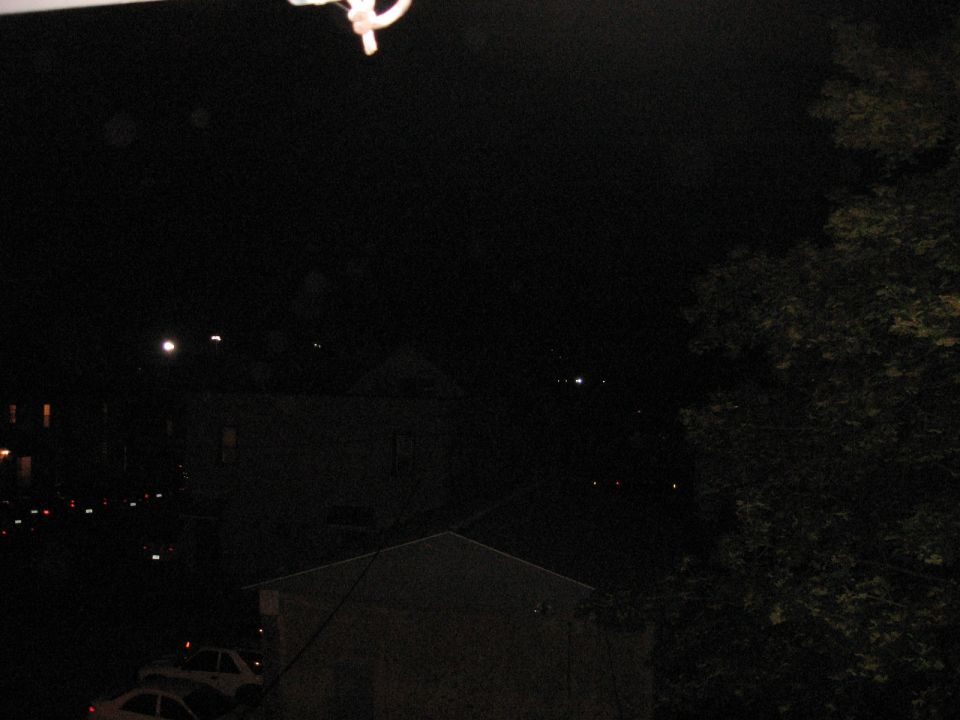 Nighttime view of houses and two cars parked on a street, with trees on the right. SUPERMOTORS.NET watermark visible