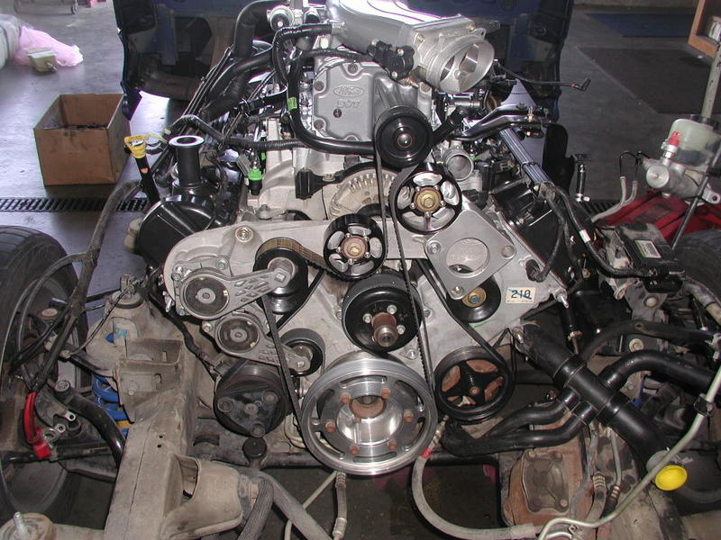 Ford V8 engine with supercharger, exposed engine bay, metal pulleys and belts, black hoses, in a workshop