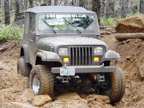1989 Ford Bronco - A gray Jeep Wrangler navigates a challenging off-road course with large rocks and mud.