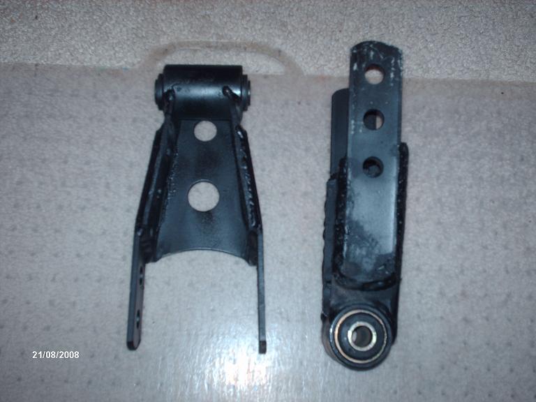 Two black metal leaf spring shackles with bushings on a textured surface. One is a triangular bracket, the other is a rectangular bracket