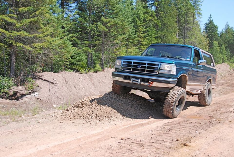 Teal Ford Bronco with lifted suspension driving on a dirt road with trees in the background