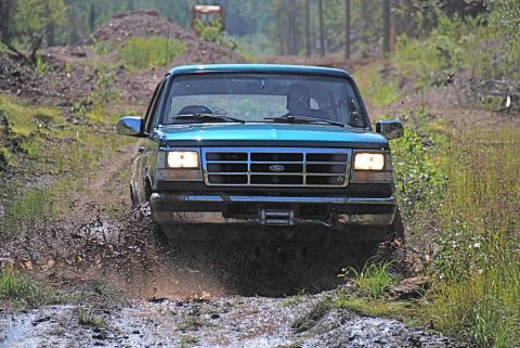1996 Ford Bronco - A teal Ford Bronco navigates a muddy trail, kicking up water and dirt.