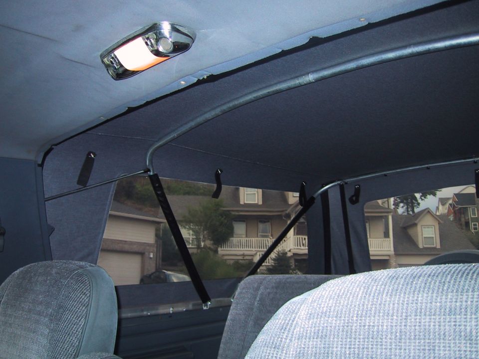 Interior shot of a vehicle, showing the headliner, dome light, and seats. Houses are visible through the windows