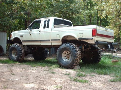 1990 Ford F-150 - A lifted, cream-colored Ford F-150 pickup truck with oversized tires sits parked on a dirt and grass lot. The truck is viewed from the rear…