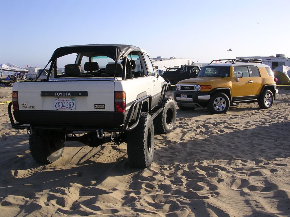 White 1985 Toyota 4Runner with soft top and off-road mods parked on sand next to yellow Toyota FJ Cruiser.