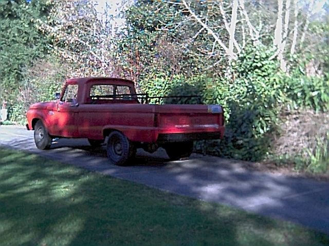 Red vintage Ford pickup truck parked on a driveway next to hedges, seen from the rear three-quarter angle