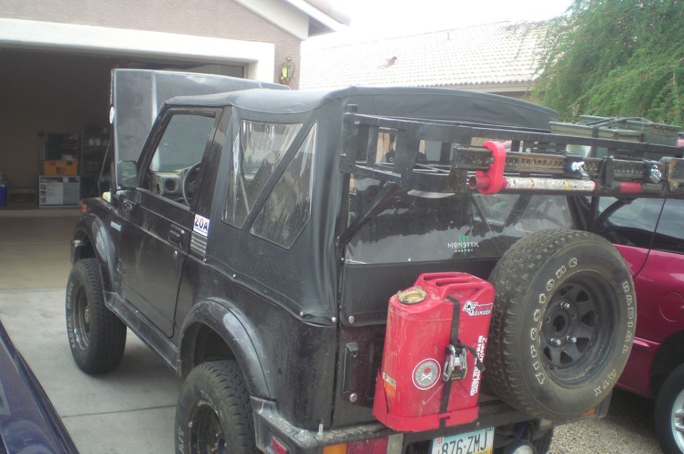 Black Suzuki Samurai with soft top, roof rack, and rear-mounted spare tire in driveway
