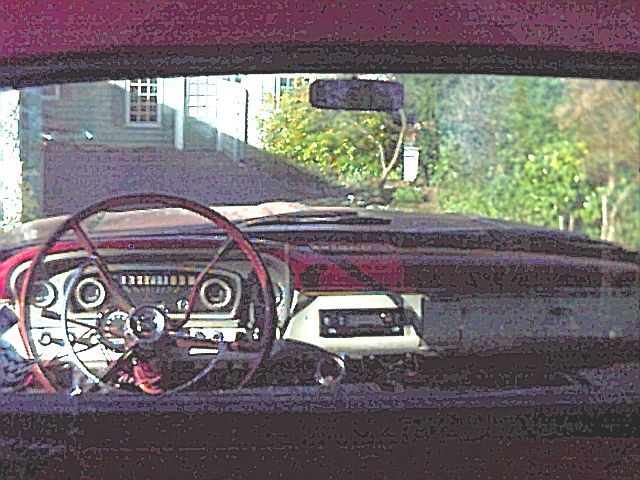 Interior of a vintage truck showing red dash, steering wheel and radio, with house and trees through the windshield