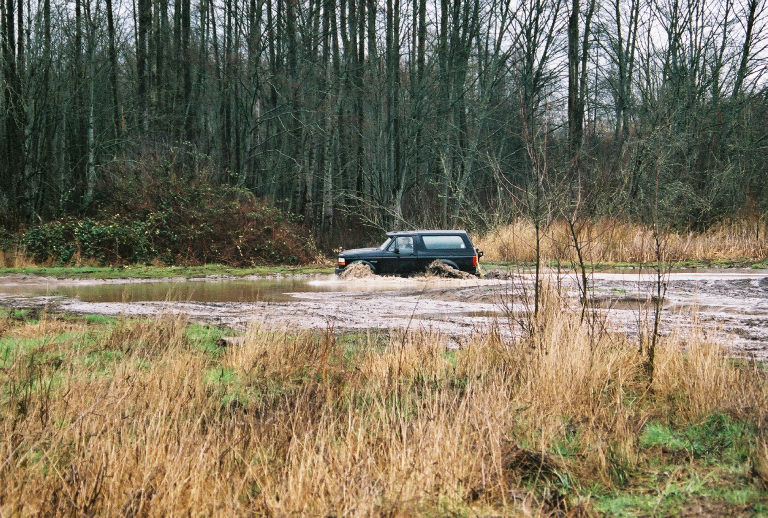 Dark SUV driving through muddy water in a wooded off-road clearing