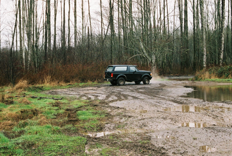 Dark green two-door Ford Bronco SUV parked on a muddy clearing in a bare-tree forest