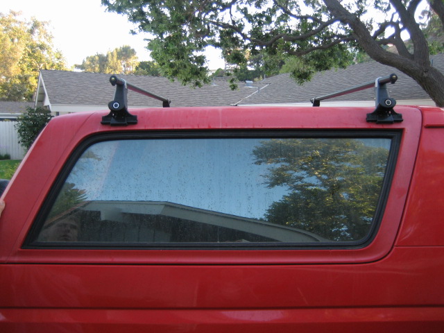 Red truck cap with roof rack, window reflecting trees and house.