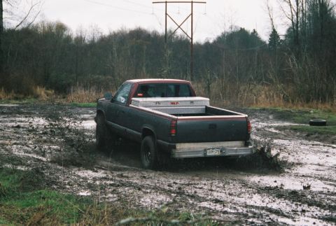 1995 Ford Bronco - A dark pickup truck with a red cab is stuck in the mud, its rear end angled towards the viewer.