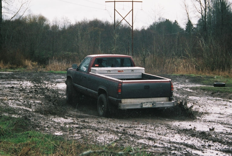 Dark pickup truck with bed toolbox driving through muddy lot, rear view