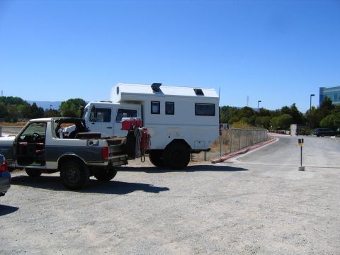 1991 Ford Bronco - A white camper shell is mounted on the back of a white truck cab, which is towing a gray and white pickup truck with its doors removed. The…