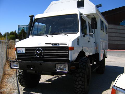 1991 Ford Bronco - A white Mercedes-Benz Unimog camper truck is parked on a gravel lot next to a chain-link fence. The truck features large off-road tires, a…