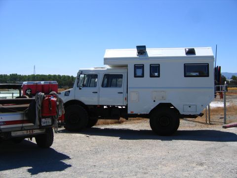 1991 Ford Bronco - A custom white expedition vehicle, likely a Unimog, is parked on a gravel lot next to a Ford pickup truck. The expedition vehicle features…