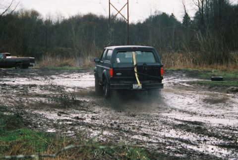 1995 Ford Bronco - Black 1995 Ford Bronco kicks up mud on a trail. A pickup truck is parked in the background.