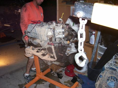 1991 Ford Bronco - A man in an orange shirt is standing next to a V8 engine that is mounted on an orange engine stand. The engine is being lifted by a black e…
