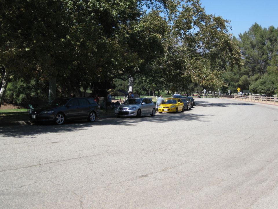 Dark gray Mazda sedan parked under trees with other cars in line.