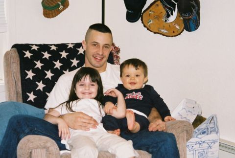 1995 Ford Bronco - A father smiles while holding his two young children on his lap, seated in a chair draped with an American flag blanket.