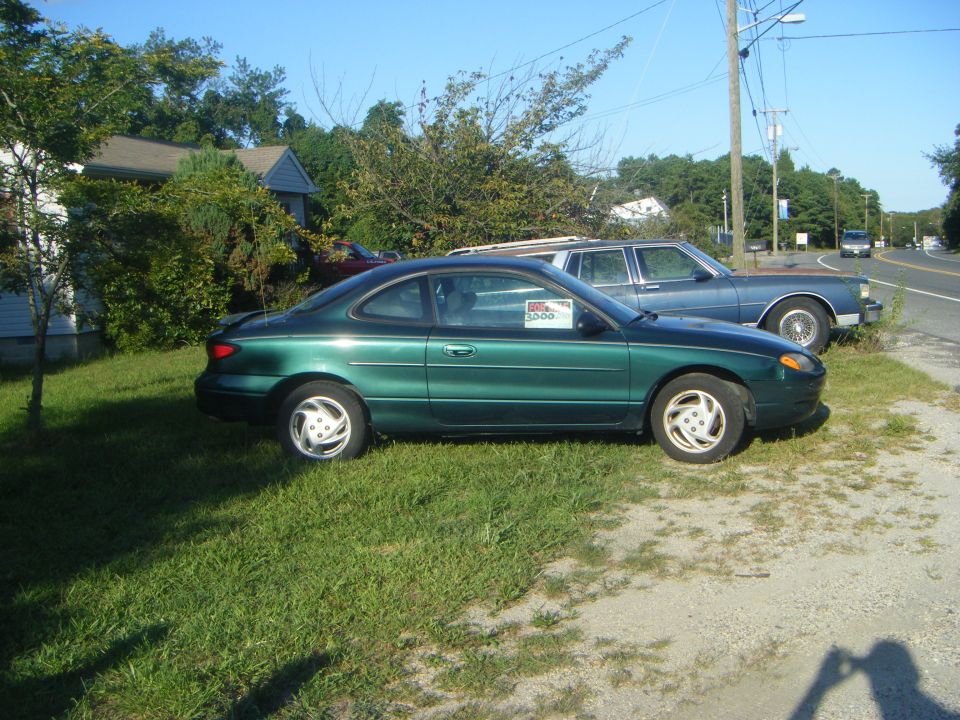 Green coupe with a "For Sale" sign in the window, parked next to a blue sedan