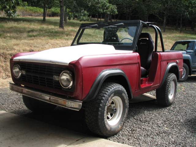 Red 1968 Ford Bronco with a white hood and black roll cage parked on gravel