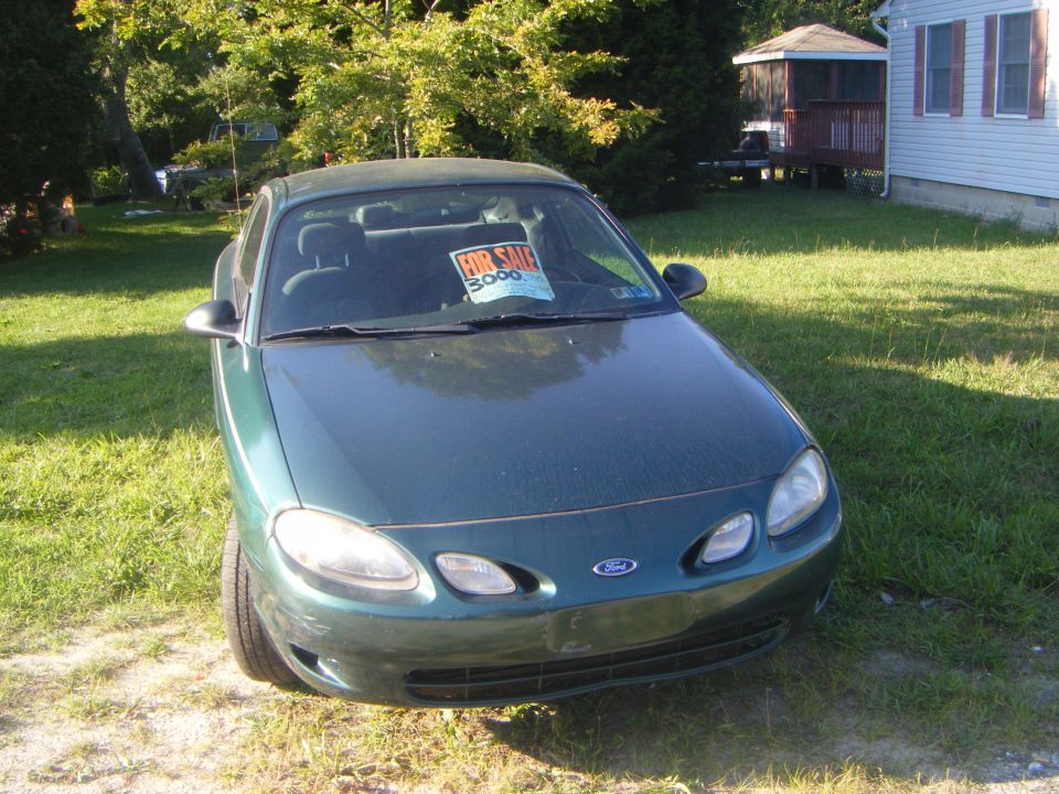 Green Ford Escort with a 'For Sale $3000' sign in the windshield parked in a grassy yard