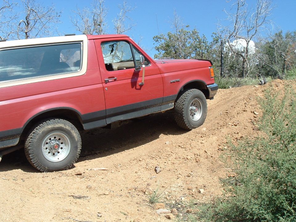 Red 1991 Ford Bronco climbing a dirt hill with trees and a clear blue sky in the background