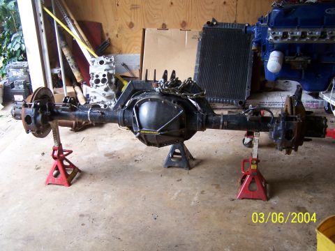 1979 Ford F-150 - Rear axle assembly, possibly from a Ford truck, is supported by jack stands in a garage setting. A blue engine block is visible in the back…