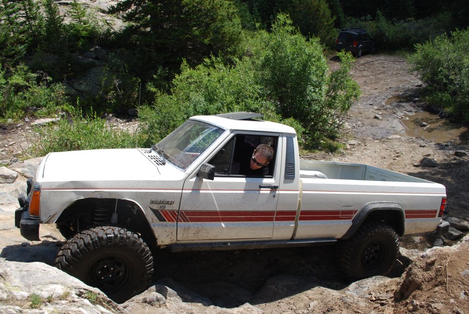 White Jeep Comanche pickup truck off-roading on a rocky trail with driver visible
