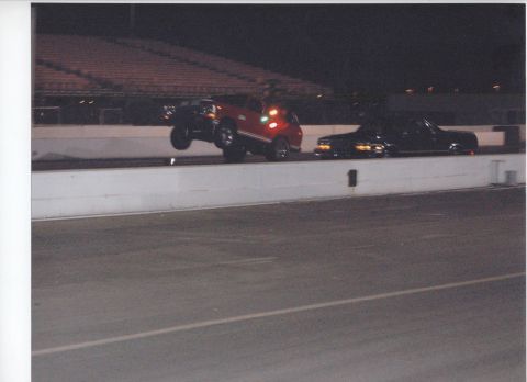 1979 Ford Bronco - A red SUV is captured mid-air, performing a wheelie on a race track at night, with another vehicle visible to the side.