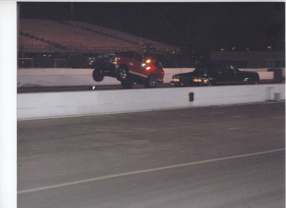 Red SUV pulling a wheelstand off the line at a night drag race beside a black car