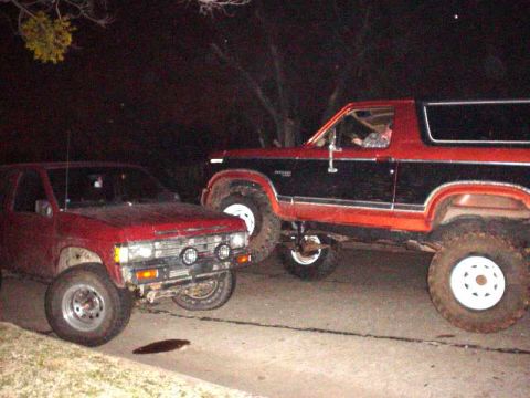 1984 Ford Bronco - Two modified off-road vehicles, a Ford Bronco and a pickup truck, are parked side-by-side at night.