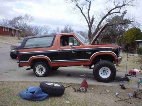 1984 Ford Bronco - A two-tone orange and black Ford Bronco is parked outside with its hood open and a spare tire nearby.