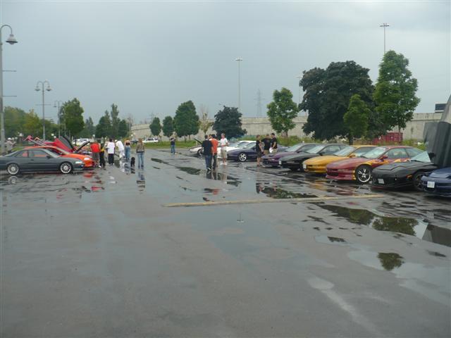 Several cars parked in a lot on a rainy day, with people standing around them