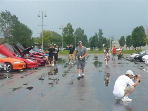 1993 Ford Probe - A gathering of cars, many with doors raised, in a wet parking lot as people walk around.