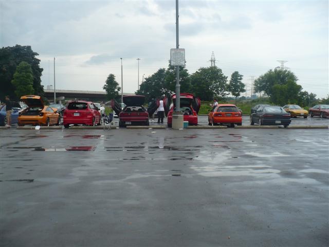 Several cars parked in a wet lot under a cloudy sky. Some car trunks are open