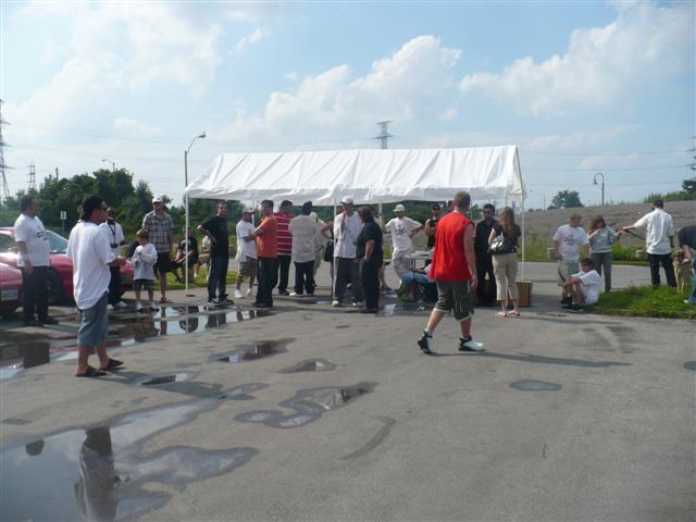 People gathered outside near a white tent. A red car is partially visible on the left