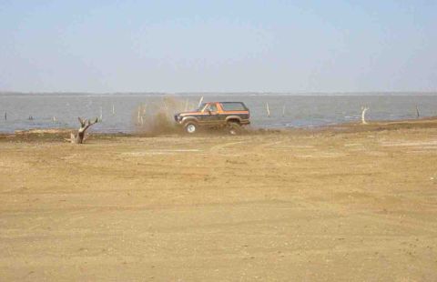 1984 Ford Bronco - A Ford Bronco drives off-road near a body of water, creating a dust cloud.