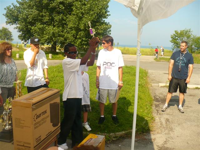 A group of people are gathered outdoors, with one person holding up a trophy. A large cardboard box with the text "DAYTEK" is visible.