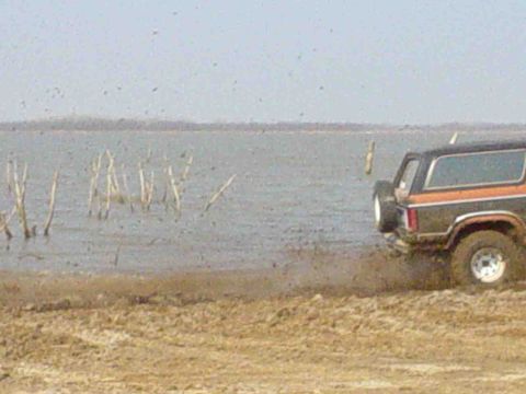 1984 Ford Bronco - SUV navigates a muddy shoreline with a body of water in the background. The Supermotors.net logo is visible in the lower right corner.