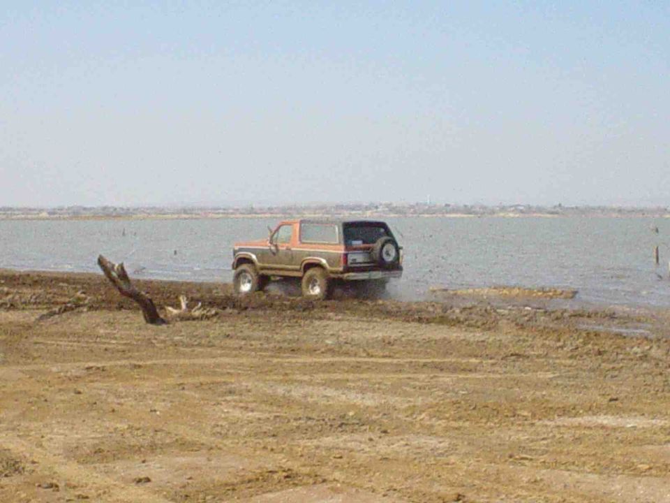 Orange and brown Ford Bronco driving into shallow water on a dirt shore